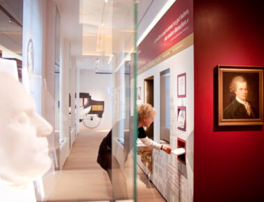 Innenansicht der Ausstellung im Lessing-Museum. Links im Vordergrund eine Gips-Totenmaske in einer Vitrine. In der Mitte eine Frau, die sich gerade Ausstellungstafeln und Bilder im Ausstellungsraum ansieht.