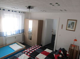 Ein Schlafzimmer mit einem Doppelbett und karierter Bettwäsche. Davor eine Einzelliege für einen weiteren Gast. Rechts an der Wand ein Kleiderschrank mit Spiegel, eine Tür und eine Kommode mit Nachtlicht. An der Wand ein Bild und ein Thermometer.. 