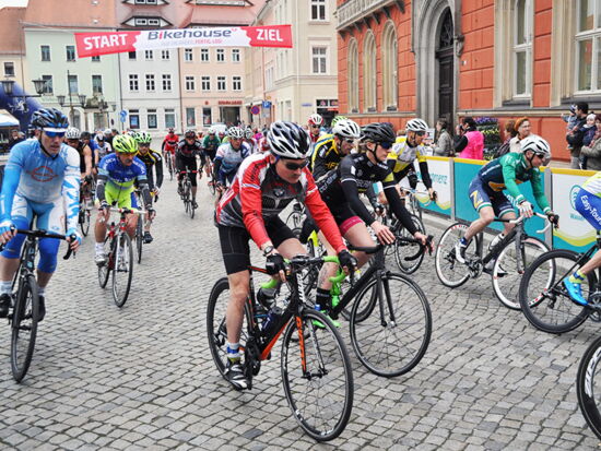 Eine große Gruppe Radfahrer zum Kamenzer Blütenlauf. Nach dem Startschuss auf dem Marktplatz ist die Gruppe in das Radrennen gestartet. Am Rand stehen Zuschauer. Ein Banner mit Aufschrift Start / Ziel ist aufgespannt.