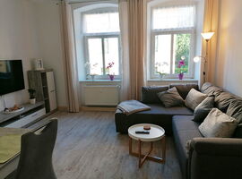 Wohnbereich mit einer Eckcouch, Stehlampe, Kissen und Decke rechts. Davor ein kleiner runder Couchtisch mit Fernbedienung und Untersetzern. Links Wohnwand mit TV und Esstisch. Im Hintergrund zwei große Fenster mit Blumen und Vorhängen.. 