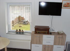 Wohnbereich der FeWo Voigt mit Kommode, Radio und TV. Links daneben ein Fenster mit Ausblick in den Gartenbereich.. 
