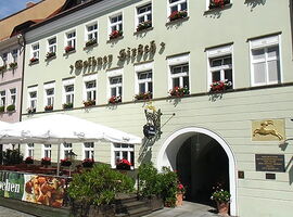 Vorderansicht des Eingangsbereiches des AKZENT Hotel Goldner Hirsch. Rechts das Goldrelief eines Hirschen. Links eine Terrasse mit Stühlen, Tischen und Schirmen.. 