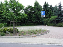 Leerer Stellplatz am Damm von der Seite / Straße aus. Im Hintergrund Bäume und Sträucher. davor die Zufahrt zum Stellplatz rechts und die drei Stellplätze links. Im Vordergrund ein P-Schild mit Wohnwagen-Symbol und ein Beet mit Blumen und einem Baum.