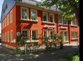 Außenansicht von Pension & Restaurant Kiautschau. Im Vordergrund links Zufahrt zum Parkplatz und Terrassenbereich mit Sitzgruppen. Rechts Eingang Pension.