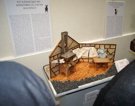Das Bild zeigt ein Exponat zur Sagenausstellung im Kamenzer Rathaus als Ausstellungsbeispiel. Es handelt sich um eine Ansicht eines Miniatur-Hauses mit kleinem Herd, Tisch und Stühlen und einer Maus, die am Tisch sitzt. Ausstellungstafeln im Hintergrund.. 