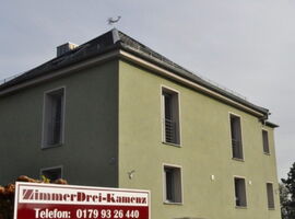 Ein grünes Wohnhaus im Stil einer modernen Villa. Zum Teil große Fenster mit Metallabgrenzung. Vor dem Wohnhaus steht ein Schild mit der Aufschrift ZimmerDrei-Kamenz, Telefon: 0179 93 26 440. 