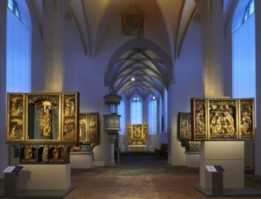 Innenansicht der Klosterkirche St. Annen / Sakralmuseum. Ausstellungsraum mit jeweils 2 beleuchteten Schnitzaltären am Rand und einem Altar im Hintergrund in der Mitte. Die Altäre veranschaulichen unter anderem Maria, Jesus und Franz von Assisi.