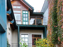 Außenansicht des Gästehaus Schillerpromenade. Ein Haus mit mindestens zwei Etagen, bemalten Holzelementen und zwei Balkonen. Rechts überranken Pflanzen die Wand.. 