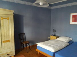 Ein Schlafzimmer mit zwei zusammengestellten Einzelbetten auf der rechten Seite. Darüber ein Bild an der Wand und ein kleiner gelber Nachttisch mit Lampe links daneben. Links ein heller Kleiderschrank und ein Holzstuhl.. 