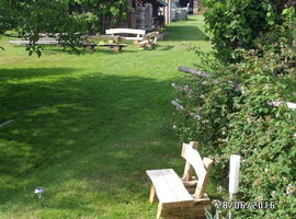 Gartenabschnitt mit Wiese. Rechts im Vordergrund ein paar Sträucher an einem Holzzaun, davor eine Holzbank. Links im Hintergrund Gebäude und eine Sitzgruppe aus drei Holzbänken. Im Vordergrund ein Baum.. 