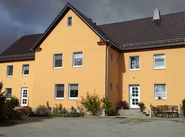 Ein gelbliches großes Wohnhaus bei Sonnenschein. Zwei Treppeneingänge mit Topfpflanzen und Blumenschmuck davor. Rechts eine Sitzgruppe mit Bank und Stühlen.. 
