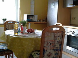 Zweites Küchenbeispiel. Rechts eine granitfarbene Küchenzeile mit Herd und Schränken. Im Hintergrund ein Regal / Bartisch mit Mikrowelle. Links steht ein ovaler Esstisch mit Tischdecke im Raum und ist umringt von vier Stühlen. Ein Obstkorb auf dem Tisch.. 