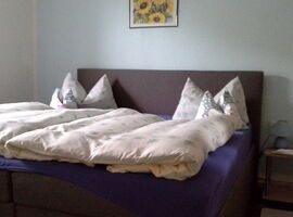 Beispiel Schlafzimmer mit einem großen Doppelbett und heller Bettwäsche. Rechts ein Nachttisch mit kleiner Leuchte. Links ein Fenster.. 