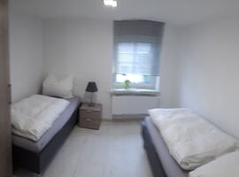 Zimmer mit je einem Einzelbett rechts und links an der Wand. Im Hintergrund ein Fenster und ein Nachttisch mit Lampe. Links im Vordergrund ein Waschbecken mit Spiegel und ein schmaler Kleiderschrank.. 
