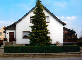 Außenansicht des Hauses Isendahl. Ein weißes Wohnhaus bei Sonnenschein mit einem Eingangsbereich mit Tfreppenaufgang links. Davor eine Steinmauer mit Hecke. Vor dem Haus ein großer Baum. Rechts ein Balkon bzw. Wintergarten.. 