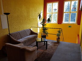 Treppenaufgang und Etage im Hostel jo-si. Auf der Etage stehen ein Sofa, ein Sessel, ein Couchtisch mit Blumen, zwei Topfpflanzen und eine Stehlampe. Im Hintergrund zwei große Fenster.. 