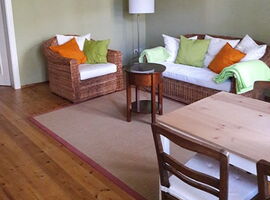 Ein Wohnraum mit grünen Wänden mit Bordüre und einem Holzfußboden mit Teppich. Rechts im Hintergrund ein Korbsessel, eine moderne Stehlampe und ein Zweier-Korbsofafa mit Kissen und Decke darauf. Davor ein kleiner runder Holztisch. Sitzgruppe rechts vorne.. 