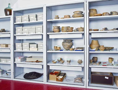 Vier hohe Regale mit Sammlungsobjekten des Museums der Westlausitz. Töpfe, Becher, Kisten, ein kleines Geweih und eine Art halber schwarzer Schild befinden sich darin. Beispiel für den Magazinbestand im Sammelsurium in Kamenz.. 