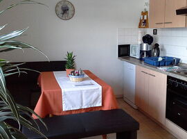 Beispiel Küche mit einer Eckbank und Esstisch auf der linken Seite. Rechts eine buchefarbene Küchenzeile mit Spüle, Herd, Kaffeemaschine, Mikrowelle, Schränken und Utensilien.. 
