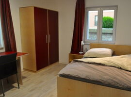 Biespielzimmer mit einem Schreibtisch und einem schwarzen Stuhle vor einem Fenster links. Daneben ein Kleiderschrank. Rechts ein Holzdoppelbett mit einem Nachttisch und Lampe vor einem weiteren Fenster. Laminatfußboden.. 