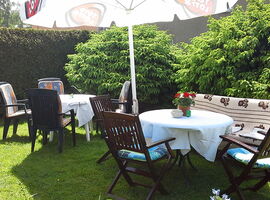 Gartenbereich mit zwei Sitzgruppen. Ein runder Gartentisch mit vier Stühlen. Ein weiterer runder Gartentisch daneben mit Blumenschmuck, drei Stühlen und einer Bank. Dabei steht ein Sonnenschirm und gibt Schatten.. 