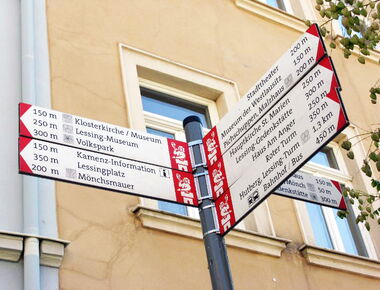 Mast mit sechs Richtungsweisungsschildern in Kamenz, die den Weg zu den verschiedenen Sehenswürdigkeiten und touristischen Anlaufstellen in der Stadt weisen, wie z.-B. zum Roten Turm, zur Kamenz-Information und zum Lessing-Museum.. 