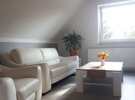 Alternative Ansicht des Wohnbereichs der FeWo am Heidelberg. Links eine weiße Ledercouch mit Beistelltisch und Palmenpflanze im Hintergrund. Sessel neben der Couch. Vor der Couch ein passender Tisch mit Läufer und Blumenschmuck. Rechts hinten ein Fenster.. 