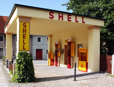 Außenansicht der historischen Schell-Tankstelle in Kamenz. Ein gelbes Gebäude mit der Aufschrift Schell und zwei rot-gelben Tankzapfsäulen im Mittelpunkt. Beispiel für das Tankstellenmuseum in Kamenz.. 