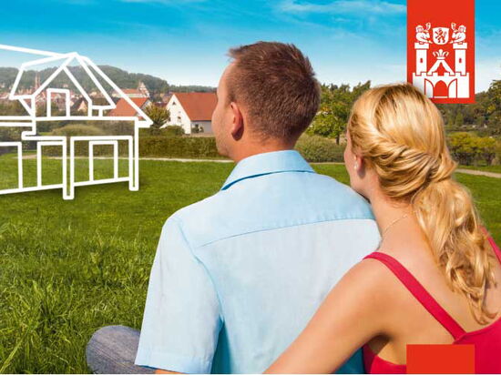 Kampagnen-Bild für das Thema Bauen. Ein Mann und eine Frau sitzen auf einer Wiese und betrachten ein illustriertes Plan-Haus auf dem Bautzner Berg - einem Baugebiet in Kamenz. Im Hintergrund weitere Häuser, grüne Wiesen und Blick auf die Altstadt.. 