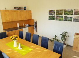 Küche und Aufenthalsraum des Hostel jo-si. Ein großer Tisch mit elf Stühlen. Im Hintergrund eine Küchenzeile mit Spüle, Geschirrspüler und Schränken und Dosen darauf. An der Wand rechts acht Naturbilder sowie zwei Topflanzen und eine Kommode.. 