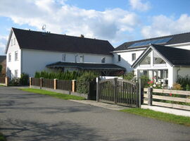 Gesamtansicht des modernen Hauses bei Sonnenschein. Grundstück ist durch Holzzäune und Doppelpforte eingegrenzt. Dazischen Gartenbereich mit Hecke, Rasen und Pflanzen. Haus mit einigen Glasfronten und Solarpanels auf dem Dach.. 