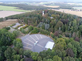 Hutbergbühne Kamenz aus der Vogelperspektive. Freilichtbühne und Konzertgelände in Form eines Amphitheaters von oben. Im Hintergrund stehen der Hutberg Aussichtsturm und die Hutberggassttätte. Dörfer in der Umgebung erkennbar.. 