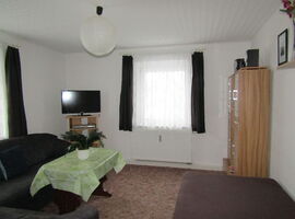 Einblick in den Wohnzimmerbereich mit einer dunklen Couch links und einem Couchtisch mit Topfpflanze davor. In der Ecke ein TV Regal mit Fernseher darauf. Rechts eine Schrankwand mit Lautsprecherboxen darauf. Der Raum wird durch zwei Fenster erhellt.. 