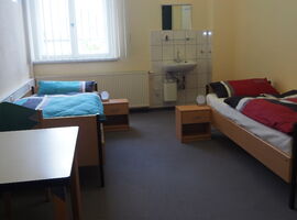 Ein Beispielzimmer im Hostel jo-si mit zwei Einzelbetten und jeweils einem Nachttisch mit Kugelleuchte. Im Vordergrund links ein Tisch. Rechts im Hintergrund ein Waschbecken mit Spiegel und Ablage.. 