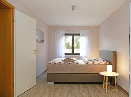 Schlafzimmer mit einem Doppelbett vor einem Fenster mit Vorhängen. Bett in Form eines Boxspringbettes mit Design-Blümchen-Bettwäsche. Rechts neben dem Bett ein Beistelltisch mit Nachttischlampe. Links ein Spiegel mit Silhouette an der Wand und eine Tür.. 