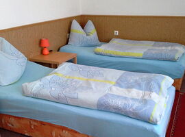 Schlafzimmer Beispiel mit zwei Einzelbetten und Blumenbettwäsche. Dazwischens teht ein hölzerner Beistelltisch mit Leuchte. An der Wand sind eine Holzverkleidung und weiße Tapete.. 