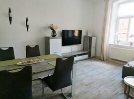 Wohn- und Essbereich der FeWo Freudenberg. Links steht ein Tisch in Granitoptik, eingedeckt mit Tischläufern und einer Dekoschale. Vier schwarze Wippstühle darum. Rechts eine Wohnwand mit TV, Fenster mit Blumen, Sofa und Couchtisch.. 