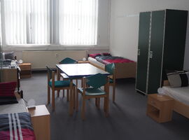 Beispielzimmer im Hostel jo-si mit vier Einzelbetten und Nachttischen mit Kugelleuchten an den Wänden. In der Mitte ein Tisch mit vier Stühlen. Rechts ein Kleiderschrank. Zwei große Fenster im Hintergrund.. 