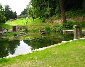Der idyllische Kamenzer Bäckerteich - ein Teich umgeben von grünen Wiesen und Bäumen im Frühling / Sommer. Teich umrandet mit Steinen und Holzzaun. Ein Entenhaus im Teich. Ein Gehweg führt links vorbei.. 