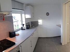 Helle moderne Küchenzeile unter einem Fenster. Links Herd mit Ofen und Abzugshaube. Nach rechts Toaster, Kaffeemaschine, Wasserkocher, Spüle und Schränke. Rechts Eingangstür.. 