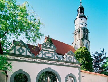 Eingangsportal der Hauptkirche St. Marien in Kamenz. Im Vordergrund ein Tor mit drei Rundbögen und der Aufschrift "Der Herr behüte deinen Ausgang und Eingang von nun an bis in Ewigkeit".. 