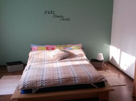Doppel-Futonbett mit unterschiedlich farbiger und gemusterter Bettwäsche. Rechts und links davon jeweils ein Läufer und Korb-Nachttische mit einer Nachttischlampe.. 