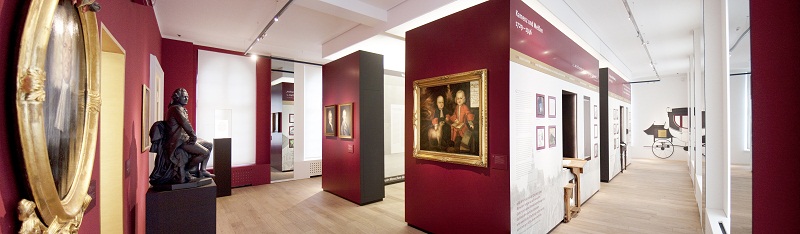 Panoramaansicht des Ausstellungsraumes im Lessing-Museum. Verschiedene Gemälde Lessings hängen an den Wänden. Links steht eine große Lessing-Skulptur. Im Mittelpunkt drei Gänge mit Ausstellungsstücken.