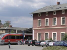 Seitenansicht des Bahnhofes in Kamenz. Eine Regionalbahn steht am Gleis. Menschen stehen davor und steigen ein bzw. aus. Auf dem Parkplatz stehen verschiedene Autos.