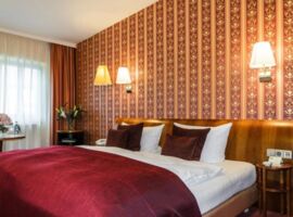 Einblick in ein Doppelzimmer des Hotels Goldner Hirsch in Kamenz. In der Mitte ein großes, gemütlich wirkendes Bett. Rechts und links stehen Nachttische mit Blumen und Kärtchen. Das Zimmer ist in edlen Rot- und Goldtönen gehalten. Übernachtungsbeispiel.