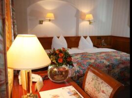 Ein Beispiel-Hotelzimmer des AKZENT Hotel Goldner Hirsch. Links im Vordergrund ein Tisch mit Broschüren, Telefon, Blumenschmuck und Lampe. Rechts im Hintergrund ein Doppelbett mit Blumen-Muster-Bettwäsche, Nachttisch und zwei Hängelampen an der Wand.