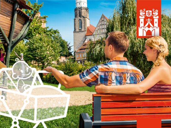 Imagebild der Kampagne Neue Stadt. Neues Glück. Ein junges Elternpaar sitzt in einem Park auf einer Bank und schaukelt einen virtuell eingezeichneten Kinderwagen links mit einem Baby darin. Im Hintergrund die Kamenzer Altstadt.