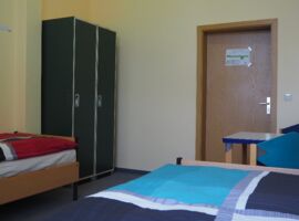 Gegenüberliegende Ansicht eines Zimmers im Hostel jo-si mit zwei Einzelbetten. Links im Hintergrund ein Kleiderschrank, rechts eine Tür und ein Tisch mit zwei Stühlen.