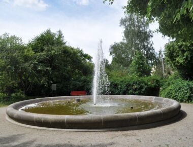 Ein Brunnen mit Wassersprenkler an der Schillerpromenade in Kamenz. Im Hintergrund steht eine rote Parkbank. Daneben ist ein Schild, das die Sehenswürdigkeit erläutert.