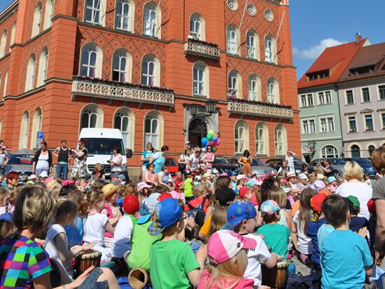 Eine große Kinder-Gruppe sitzt auf dem Marktplatz vor dem Kamenzer Rathaus. Viele von ihnen haben eine Handtrommel vor sich. Sie trommeln gemeinsam zu einer Kinderveranstaltung. Im Hintergrund das Rathaus, Luftballons, Autos und Eltern bei Sonnenschein.
